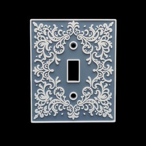May include: A decorative light switch cover with a blue-gray background and a white, ornate floral design. The cover has a rectangular shape with a single switch opening and two screw holes. The design is symmetrical and detailed.