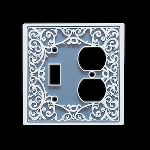 May include: A decorative light switch cover with a white, ornate floral design. The cover has a light blue center with cutouts for a toggle switch and two electrical outlets. The design is square and sits against a black background.