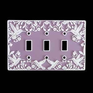 Pode incluir: Uma placa decorativa para interruptores de luz com um centro lavanda e uma borda branca ornamentada. A borda apresenta desenhos de beija-flores e florais. A placa tem três aberturas para interruptores e três orifícios para parafusos.