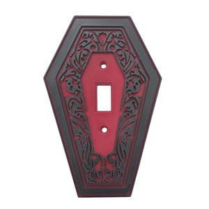 Può includere: Una copertura decorativa per interruttore della luce a forma di bara. La copertura è nera con un centro rosso e presenta un design ornato e vorticoso. L'apertura dell'interruttore è rettangolare e sono presenti due fori per le viti.
