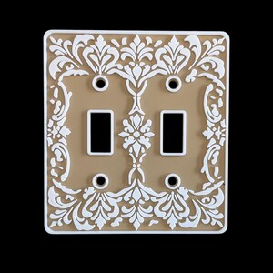 May include: A decorative double light switch cover with a beige background and white floral design. The cover has two rectangular switch openings and four screw holes. The design is symmetrical and ornate, with a white border.