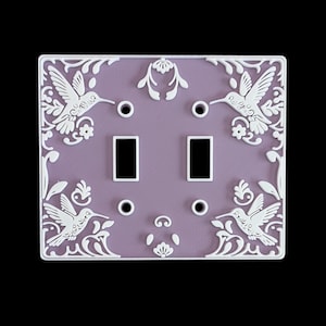 May include: A decorative light switch plate with a lavender background and white hummingbird and floral designs. The plate has two toggle switch openings and four screw holes. The frame is white and features detailed relief patterns.