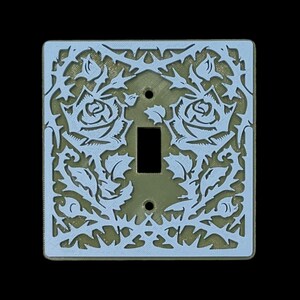 Könnte beinhalten: Dekorative Lichtschalterabdeckung mit floralem Rosendesign. Die quadratische Abdeckung hat eine rechteckige Öffnung für den Schalter. Das Design ist hellblau auf goldenem Hintergrund.