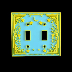 Może przedstawiać: Dekoracyjna osłona włącznika światła o tematyce niebiańskiej. Osłona jest jasnoniebieska z żółtymi akcentami, w tym gwiazdami, półksiężycem i ozdobnymi wzorami narożników. Posiada dwa prostokątne otwory na przełączniki i dwa zestawy okrągłych otworów.