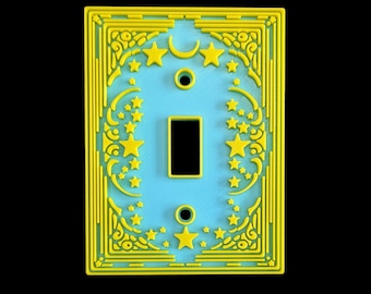 Op de afbeelding: Een decoratieve lichtschakelaar met een hemels ontwerp. De afdekking heeft een lichtblauwe achtergrond met gele sterren, een maan en sierlijke details. De schakelaaropening is zwart.