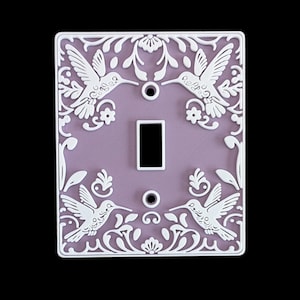 Può includere: Copri interruttore decorativo con sfondo lavanda e design floreale e colibrì bianco. Il coperchio ha un'apertura rettangolare per l'interruttore e due fori per le viti. Il design del colibrì è ripetuto quattro volte.