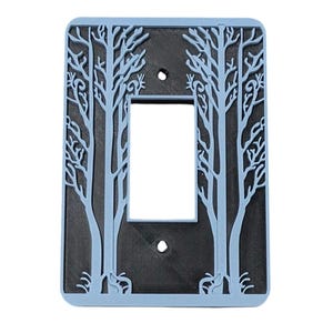 Puede incluir: Tapa de interruptor de luz de plástico azul claro con un diseño recortado de dos árboles. Los árboles tienen un fondo gris oscuro.