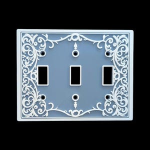 Puede incluir: Una placa decorativa para interruptores con un fondo azul claro y un diseño floral blanco ornamentado. La placa tiene tres aberturas rectangulares para interruptores y un marco rectangular con un patrón detallado.