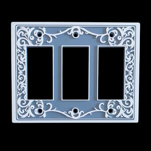 May include: A decorative light switch plate with three openings. The plate has a light blue centre and a white, ornate, floral design around the edges. The design is symmetrical and adds a touch of elegance.