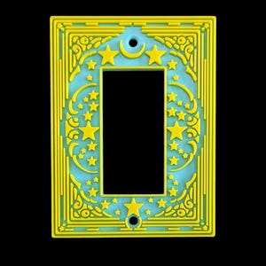 Puede incluir: Una cubierta decorativa para interruptor de luz en amarillo y azul claro. La cubierta tiene forma rectangular con una abertura central, adornada con estrellas, una luna creciente y adornos ornamentados. El diseño es simétrico y detallado.