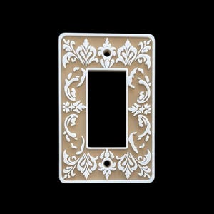 May include: A decorative light switch cover with a beige background and white floral scrollwork design. The cover has a rectangular shape with a single switch opening and is set against a black background.