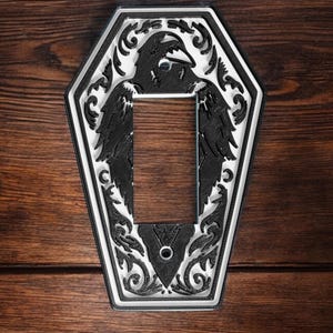 Pode incluir: Tampa de interruptor de luz decorativa em preto e branco com um design de corvo. A tampa tem formato de caixão e apresenta uma ilustração detalhada de um corvo com volutas ornamentadas. O design está em uma borda branca.
