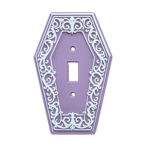 May include: A lavender-coloured light switch cover shaped like a coffin. It features a white, ornate scrollwork design around the edges and switch opening. The cover has two screw holes.