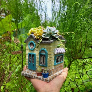 Pode incluir: Um vaso em miniatura em forma de casa com fachada de tijolo amarelo, portas e molduras de janelas turquesa e um toldo listrado. O telhado é coberto com suculentas. Uma placa que diz "Shalom" está presa a um poste.