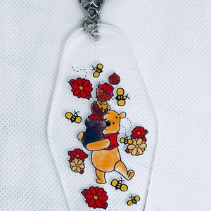 May include: Clear acrylic keychain featuring a cartoon bear carrying a honey pot. The keychain has a silver keyring and chain. The bear is surrounded by red flowers, bees, and honey pots. A charming accessory for fans of the beloved character.