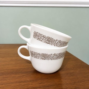 May include: Two white ceramic cups stacked vertically, each featuring a brown line-art floral pattern encircling the middle. The cups have a rounded body and a single handle on the side, showcasing a consistent botanical design with leaves and flowers.