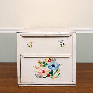 May include: A white metal storage cabinet with two drawers, featuring hand-painted floral motifs in blue, pink, green, yellow, and red. The top drawer displays small floral accents, while the bottom drawer features a larger, central bouquet design.