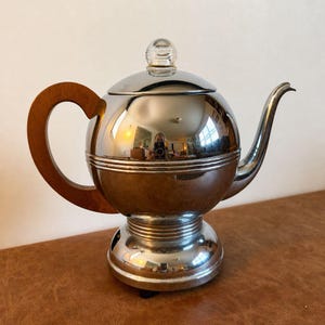 May include: A polished chrome teapot with a spherical body, a curved wooden handle, and a matching chrome lid topped with a clear glass knob. The teapot features a long, slender spout and a tiered, ribbed base design with a reflective finish.