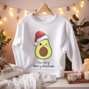 Könnte beinhalten: Weißes Sweatshirt mit einem Cartoon-Avocado, die einen Weihnachtsmannhut trägt. Die Avocado hat ein lächelndes Gesicht und einen braunen Kern. Der Text "Avo Very Merry Christmas" ist darunter aufgedruckt. Das Sweatshirt hängt an einem Holzbügel.