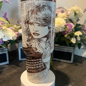 May include: A brown and white stainless steel tumbler with a vintage typewriter and a woman's face design. The tumbler has a white lid and is sitting on a white base.