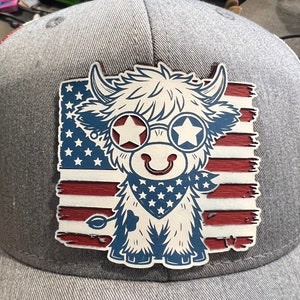 Puede incluir: Una gorra de béisbol gris con una vaca de dibujos animados blanca y peluda que lleva gafas de sol y un pañuelo con estrellas y rayas. La vaca está de pie frente a una bandera estadounidense.