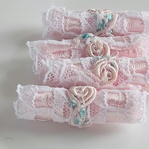 May include: Close-up of four stacked, decorative fabric rolls. Each roll is wrapped in pink satin and white lace, with a small, decorative embellishment in the center. The embellishments are a light pink color with a touch of blue.