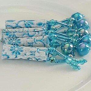 May include: A collection of decorative blue and white paper wrapped items with beaded accents. The paper is white with a blue floral design. The beaded accents are a light blue color and have a variety of shapes and sizes.