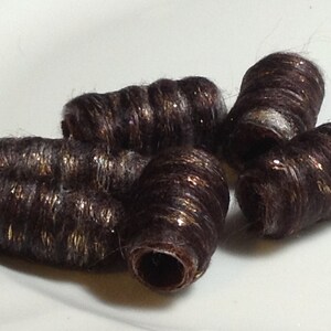 May include: A collection of dark brown, cylindrical hair beads with gold flecks. The beads are arranged on a white surface, showcasing their textured, rolled appearance. These decorative hair accessories add a touch of sparkle.