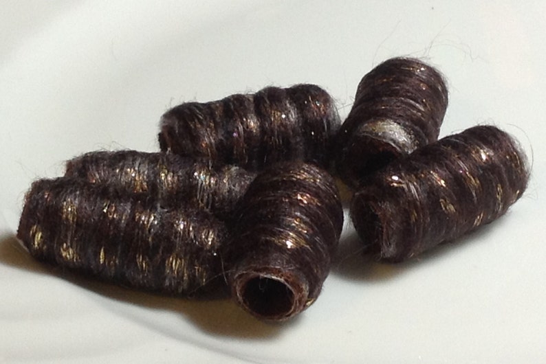 May include: A collection of dark brown, cylindrical hair beads with gold flecks. The beads are arranged on a white surface, showcasing their textured, rolled appearance. These decorative hair accessories add a touch of sparkle.
