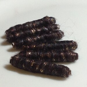 May include: Several dark brown hair extensions with gold highlights are arranged on a white surface. The extensions are cylindrical and appear to be made of synthetic hair. The gold highlights add a touch of sparkle.