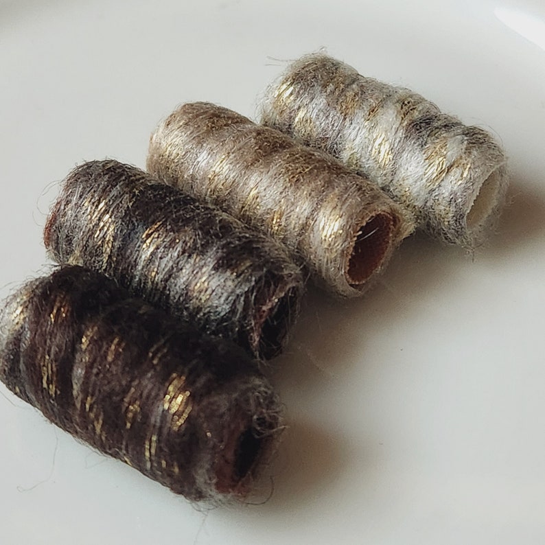May include: Four cylindrical, decorative beads in shades of brown, tan, and gray, with gold metallic accents. The beads are arranged diagonally, showcasing their textured, rolled design. The ends of the beads reveal a hollow interior.