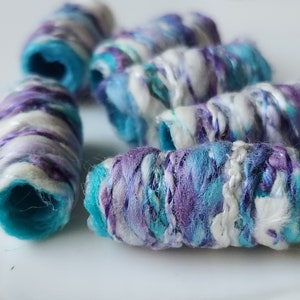 May include: A close-up of several colorful, hand-wrapped beads made of fabric scraps in shades of blue, white, and purple. The beads are arranged in a row on a white surface.