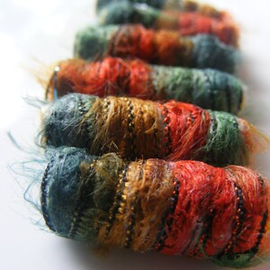 May include: A close-up of a row of colorful, hand-crafted yarn wrapped ornaments. The ornaments are made with a variety of textures and colors, including teal, brown, gold, and red.