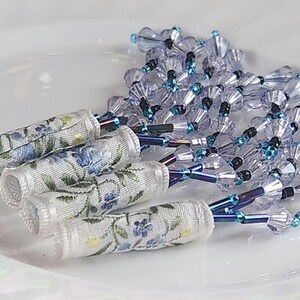 May include: Close-up of handmade floral beaded jewelry. The design features clear crystal beads with blue accents, and fabric tubes with blue floral embroidery. The jewelry is arranged on a white surface.
