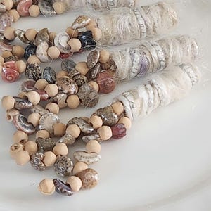 May include: A necklace made of small seashells and wooden beads. The necklace is white, brown, and pink. The necklace is on a white surface.