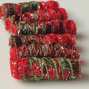 May include: Close-up of several red, green, and brown fabric tubes. The tubes are made of a textured, woven material with metallic accents. The tubes are stacked on top of each other.