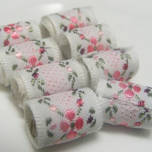 May include: A close-up of a set of white fabric trim with pink floral designs. The trim is rolled into small circles and arranged in a cluster.