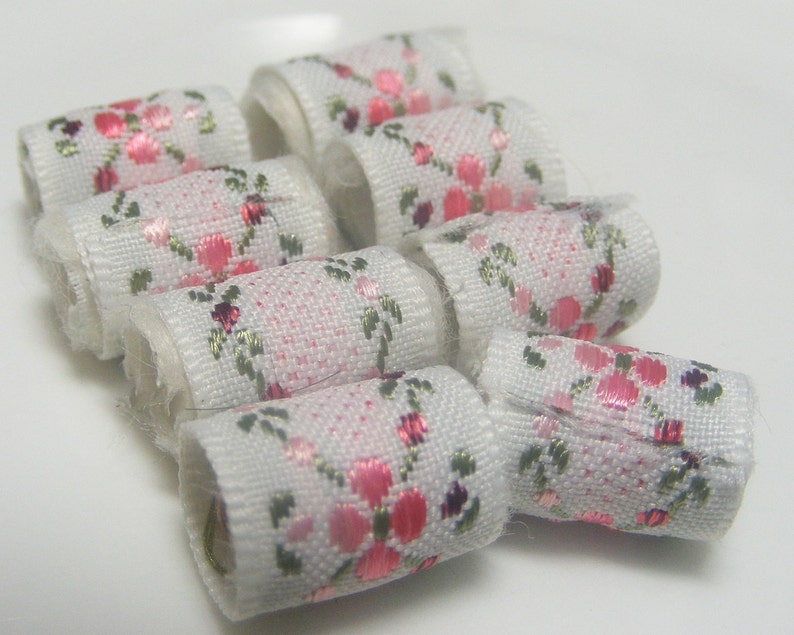 May include: A close-up of a set of white fabric trim with pink floral designs. The trim is rolled into small circles and arranged in a cluster.