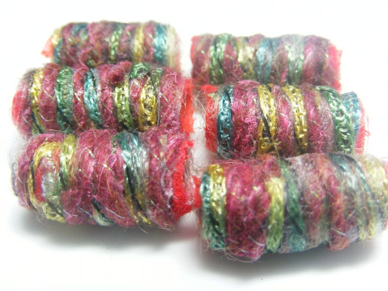 Op de afbeelding: Close-up van zes kleurrijke garen kralen. De cilindrische kralen zijn voornamelijk diep roze, met gouden, groene en turquoise accenten. Het garen heeft een getextureerde uitstraling, met metallic draden erin geweven. De kralen zijn op een witte ondergrond geplaatst.