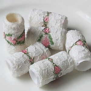 May include: Six white lace trim rolls with pink and green floral embroidery. The trim is rolled up and the ends are visible.