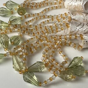 May include: Close-up of decorative beaded tassels. The tassels feature clear and gold beads, with olive green teardrop-shaped crystals. The top of the tassels are wrapped in a silver and white patterned fabric. These tassels are ideal for craft projects.