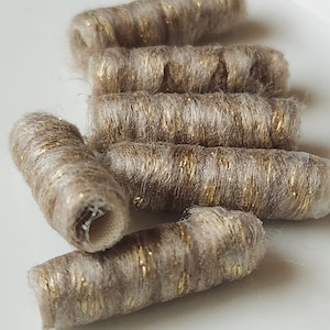 May include: Close-up of several cylindrical, beige-colored yarn tubes with gold metallic accents. The tubes are arranged on a white surface, with some in focus and others slightly blurred, creating a textured appearance.