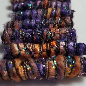 May include: A close-up of a pile of purple and orange fabric yarn with black and gold accents. The yarn is twisted and has a sparkly finish.