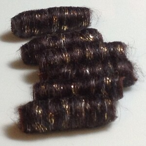May include: Several dark brown hair extensions with gold glitter accents are arranged on a white surface. The extensions are cylindrical in shape and appear to be made of synthetic hair. The glitter adds a touch of sparkle.
