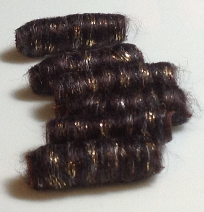 May include: Several dark brown hair extensions with gold glitter accents are arranged on a white surface. The extensions are cylindrical in shape and appear to be made of synthetic hair. The glitter adds a touch of sparkle.