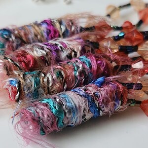 May include: Close-up of colorful, handmade jewelry featuring strands of pink, blue, and turquoise thread wrapped around a core, accented with silver and black thread. The jewelry is adorned with small, faceted, orange-red gemstones.