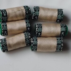 May include: Six beige fabric spools with decorative green and black patterned trim. The spools are cylindrical and arranged on a white surface. The trim features a detailed design, adding a touch of elegance to the fabric.