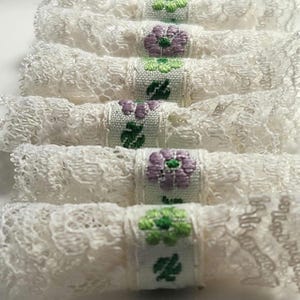 May include: Rolled white lace napkins with embroidered floral bands in green and purple. The napkins are arranged in a row, showcasing the delicate lace texture and the detailed floral embroidery.