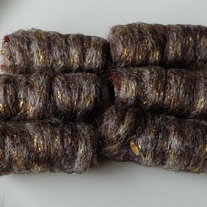 May include: Close-up of seven cylindrical yarn bundles. The yarn is a mix of brown and gray fibers with gold metallic threads woven throughout. The ends of the bundles have a reddish hue, and they are arranged on a white surface.