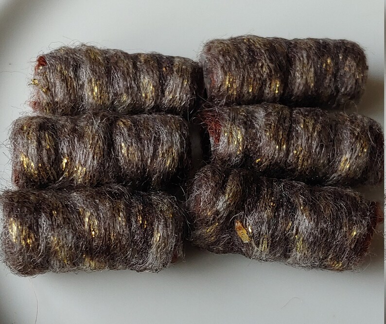 May include: Close-up of seven cylindrical yarn bundles. The yarn is a mix of brown and gray fibers with gold metallic threads woven throughout. The ends of the bundles have a reddish hue, and they are arranged on a white surface.